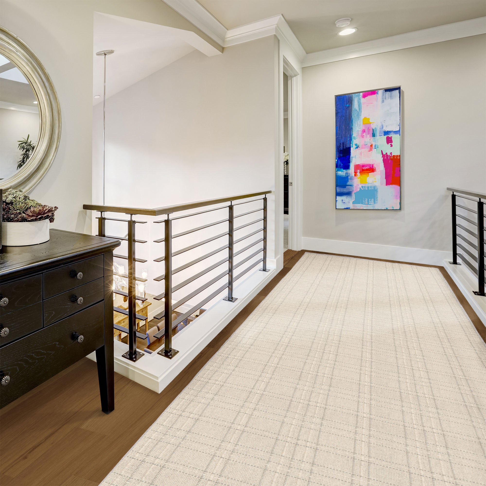 A long cream-colored area rug with a subtle grid pattern is displayed on a wooden staircase landing. The hallway features a dark wood console table on the left, a modern metal railing, and a colorful abstract painting on the wall.