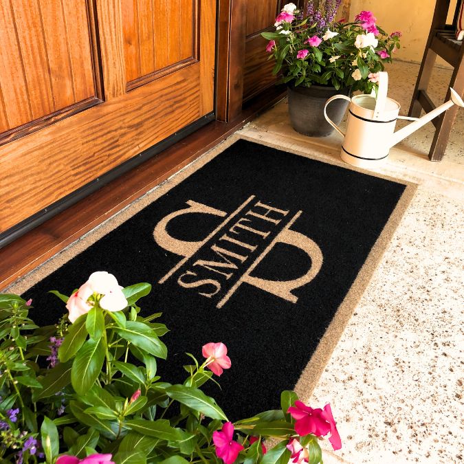 Custom black doormat with tan border and large monogram design reading 'Smith' in front of wooden door, surrounded by pink flowers and a white watering can